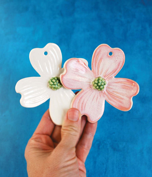 Hand holding two ceramic ornaments in the shape of dogwood flowers. One is white and one is pink.