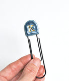 A hand holding a black metal paperclip with a dark green ceramic top featuring a gold letter 'K' on a plain white background.