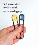 A hand holding two small decorative paperclip bookmarks with rounded tops, one gold with a yellow letter D and one blue with a blue letter K, against a white background with text that says 'Order more than one bookmark to save on shipping.'.
