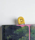 A small gold metal clip with a raised letter 'D' on a dark book or box with pink paint dripping down the side, against a plain white wall.