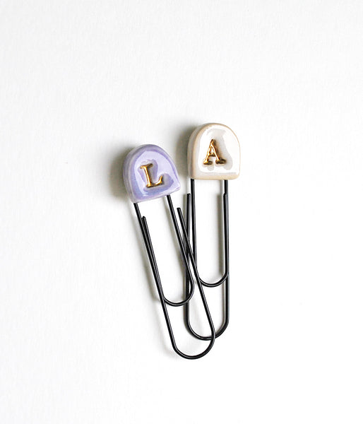 Two paperclip-style letter initial bookmarks or clips on a white background. One has a lavender top with a gold 'L', and the other has a cream top with a gold 'A'.