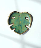 Handmade ceramic monstera-leaf shaped wall hanging. In dark green glaze, with gold luster around the edges. 